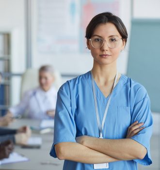 Waist up portrait of young nurse wearing glasses standing with arms crossed and looking at camera against medical conference background, copy space